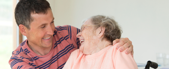 Young man has his arm over the shoulder of an elderly woman in a wheelchair as she laughs