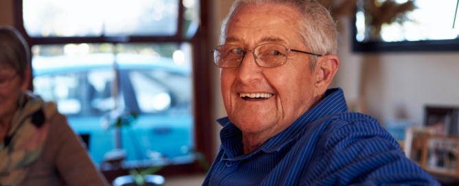 A smiling senior man, happy as result of successful long-distance caregiving, is sitting at a table and looking over his shoulder.