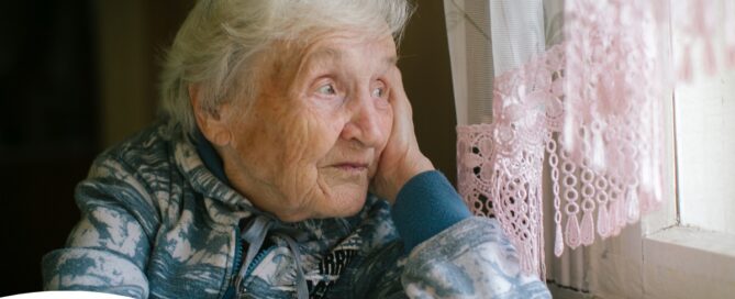 An older woman peacefully looks out of the window while the sun is still up, representing what can happen with sundowning.